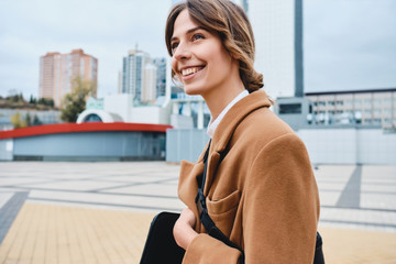 Young cheerful stylish woman in coat happily looking away while walking through city street