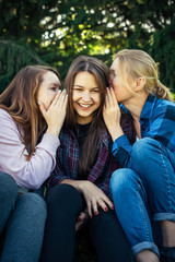 Three cheerful girls whisper and gossip against green foliage in the park. Women joke and laugh,...
