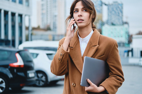 Young pensive businesswoman in coat with laptop talking on cellphone while intently looking away on city street