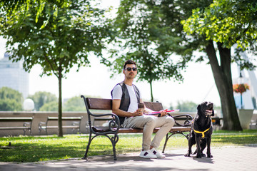 Young blind man with white cane and guide dog sitting in park in city.