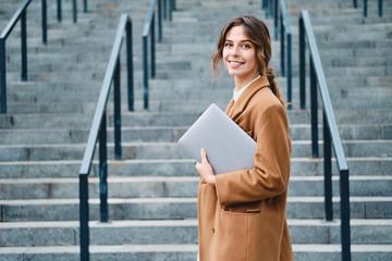 Young pretty businesswoman in coat with laptop happily looking in camera outdoor