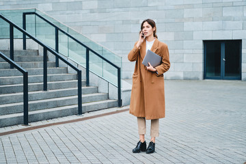 Young attractive businesswoman in coat with laptop confidently talking on cellphone on street