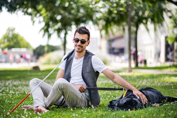Young blind man with white cane and guide dog sitting in park in city.