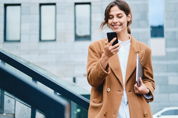 Young pretty businesswoman in coat with laptop joyfully using cellphone outdoor