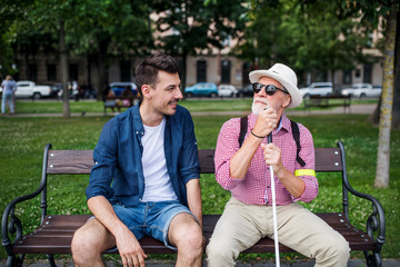 Young man and blind senior with white cane sitting on bench in park in city.