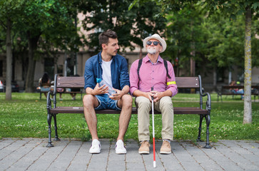 Young man and blind senior with white cane sitting on bench in park in city.