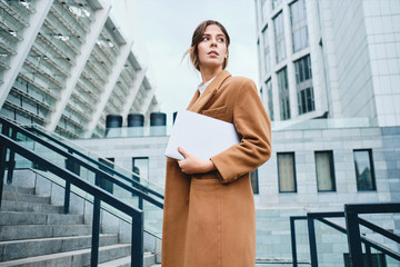 Young stylish businesswoman in coat with laptop thoughtfully looking away on street