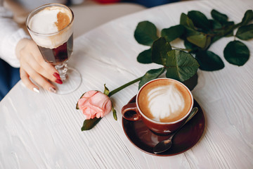 Female hand is holding a dessert. Also, there is rose and cappuchino on a table. The table is wooden.