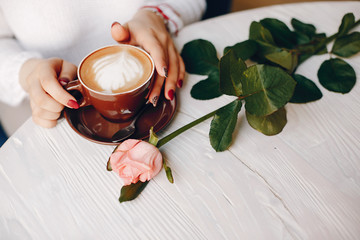 Female hands are holding a cup of coffee. There is rose on a table. The table is wooden.