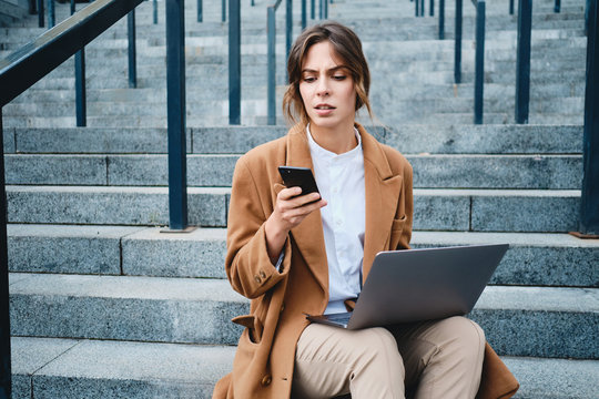 Young serious businesswoman in coat discontentedly using cellphone working with laptop on stairs on city street