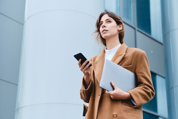 Young serious businesswoman in coat with laptop and cellphone intently looking away outdoor