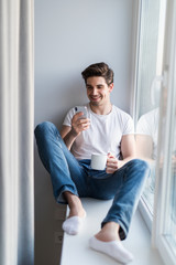 Young handsome man use phone while drinking coffee on windowsill