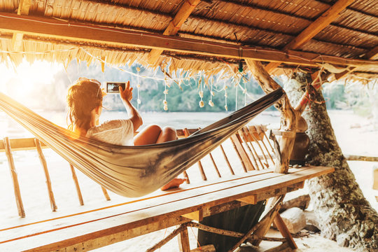 Young female lying in hammock and taking a landscape photo using modern smartphone on Samui island in Thailand. Careless summertime exotic vacation concept image.