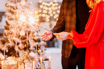 New Year 2020. New Year mood. Table with a gold tablecloth, decorated with candles and tableware for celebration. Festive still life by candlelight. A young couple lights sparklers. Close-up.