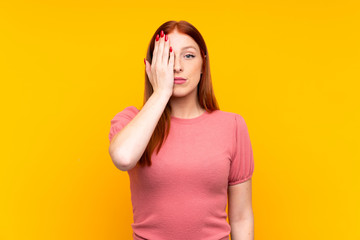 Young redhead woman over isolated yellow background covering a eye by hand