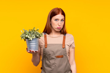 Young redhead gardener woman holding a plant over isolated yellow background sad