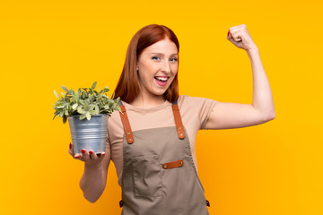 Young redhead gardener woman holding a plant over isolated yellow background making strong gesture