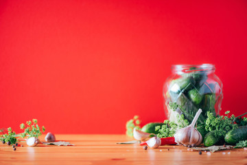 Pickling cucumbers in cans. Ingredients for marinated gherkins, glass jar, garlic, dill, salt, pepper on red background. Copy space. Concept of vegetables preservation.