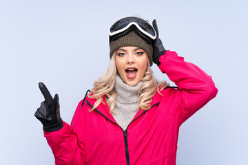 Skier teenager girl with snowboarding glasses over isolated blue background surprised and pointing finger to the side