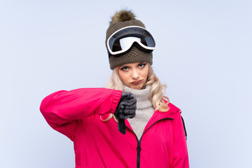 Skier teenager girl with snowboarding glasses over isolated blue background showing thumb down sign