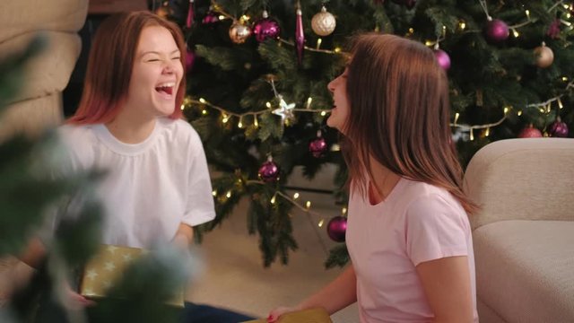 Christmas party. Home celebration. Two cheerful sisters exchanging gifts sitting on floor at home.