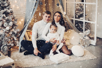 Stock photo portrait of happy family with little daughter sitting on cozy carpet under canopy decorated with garland and illumination. They are smiling at camera snuggling together. © Вячеслав Косько