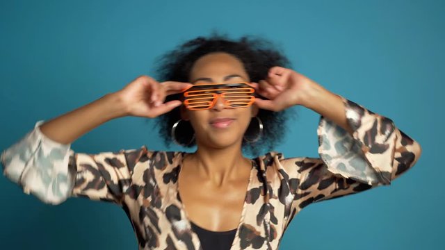 Portrait of girl with curly afro hair in neon glasses, she dancing comically with delight. Surprised excited happy woman winning. Mixed race female shocked model on blue background.