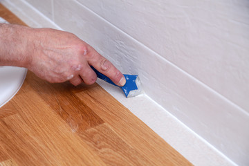Closeup hands of professional plumber worker applying white sealant, joint compound, caulk to joint of wooden table top, beige tiled wall with rectangular tile using blue scraper - obrazy, fototapety, plakaty