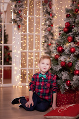 Boy in a checkered shirt and tie butterfly sitting near by Christmas tree.