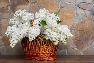 A rattan basket with a bouquet of white lilacs and a gray mouse, a stone wall in the background. Front view, place for text, Copy space.