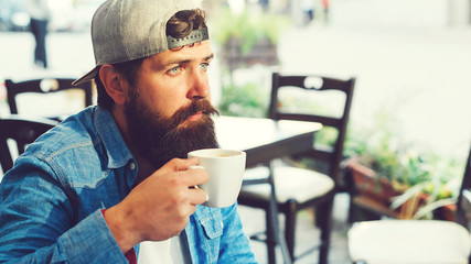 Handsome bearded guy drinks coffee at cafeteria outdoors. Man thinking and planning day. Hipster has break time