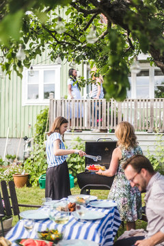 Daughter and mother preparing barbecue food while smiling man sitting at table in backyard during weekend