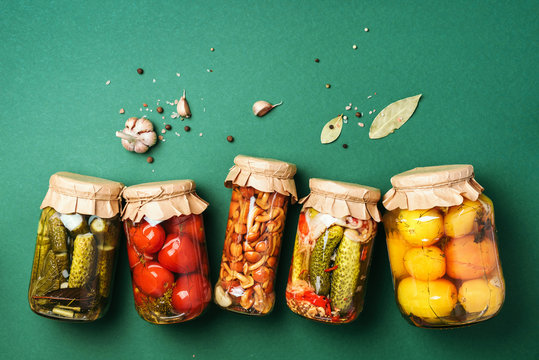 Canned and preserved vegetables in glass jars over green background. Top view. Flat lay. Copy space.