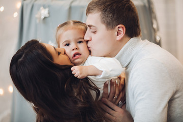 Stock portrait of attractive Caucasian family hugging and smiling at camera with daughter on hands. Brunette mother and father with adorable daughter smiling at camera.