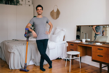 Portrait of confident mid adult man with vacuum cleaner standing in bedroom