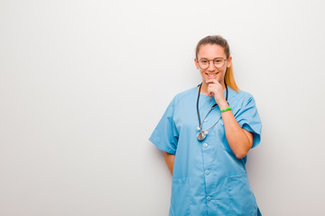 young latin nurse looking happy and smiling with hand on chin, wondering or asking a question,...