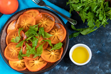 Obraz na płótnie Canvas Persimmon Salad. Thinly sliced fruit slices lie on a brown plate, sprinkled with arugula leaves. Vegetarian food. Copy space. 