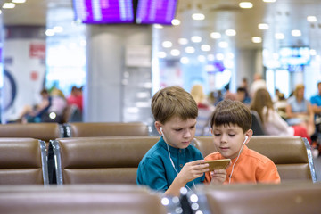 Two little boy sitting in an airport departure hall contentedly playing on his tablet or mobile phone as they wait for his flight with his luggage. Children are laughing and use earphones.