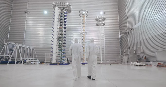 Survey in fundamental research center. Two scientiscts in protective clothes walk through high voltage testing laboratory