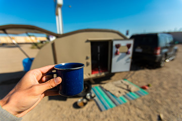 Selective focus of person holding a cup of coffee or tea in front of a parked car and an opened caravan 