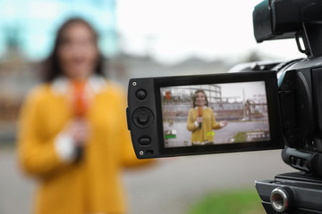 Young female journalist with microphone working on city street, focus on camera display
