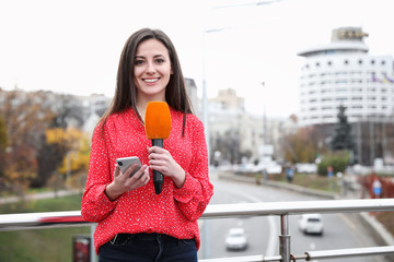 Young female journalist with microphone and smartphone working on city street. Space for text