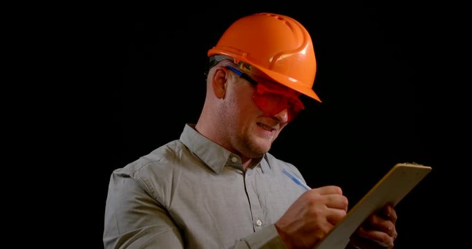 portrait of a cheerful man in a gray shirt and orange construction helmet and goggles. he writes something on a tablet, looks at the camera, smiles and continues to make notes. black background