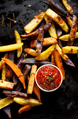 French fries,  baked fries from different types and colors of potatoes sprinkled with herbs and spices with spicy tomato sauce on a black background, top view