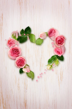 Pink rose flowers, buds and glitter confetti in a heart shape arrangement on white wooden background