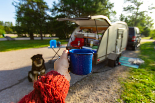 Selective focus of a person holding a cup of coffee or tea with in the background an open caravan and a dog 
