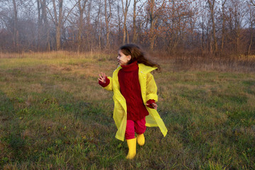 Girl child in a yellow raincoat runs in the rain in front of a coniferous forest on a rainy, foggy day. Concept of lifestyle Travel outdoor Recreation.Child looks deep into the forest in the distance.