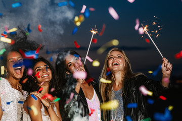 Four happy girls celebrating at night under colorful confetti holding sparkles and laughing