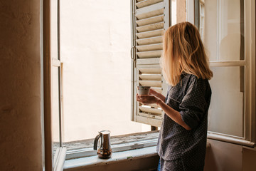 Woman in pajamas drinks morning coffee near window with shutters. Beginning of a new day. Hot drink...