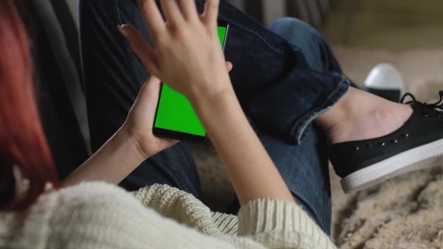 girl Gesture flips through photos with fingers using your phone with green screen. chromakey smartphone. woman in jeans resting on couch and holding smartphone. Use green mockup to insert images.